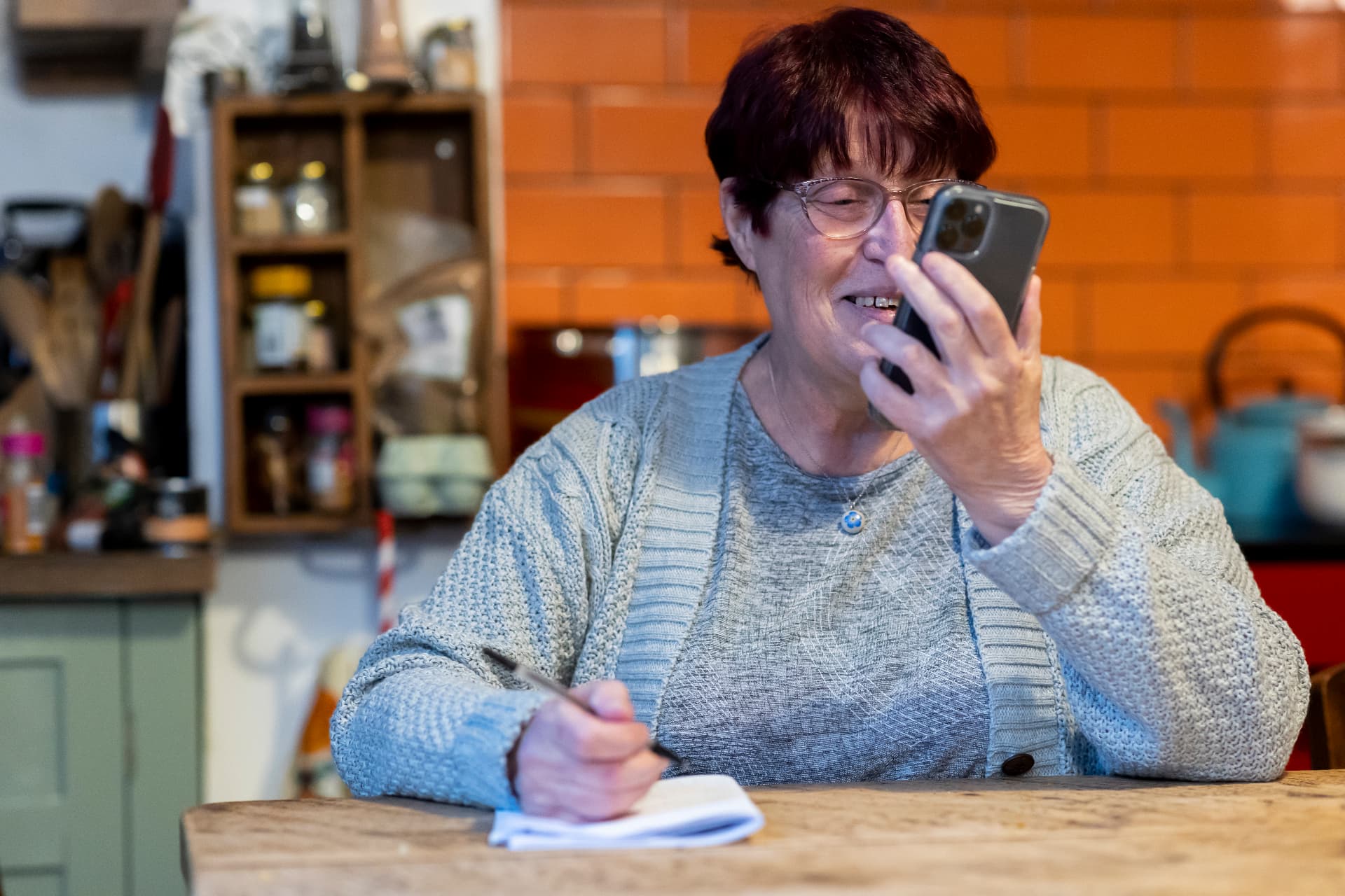 An older woman at her kitchen table making notes about the cost of home support as she talks on the phone