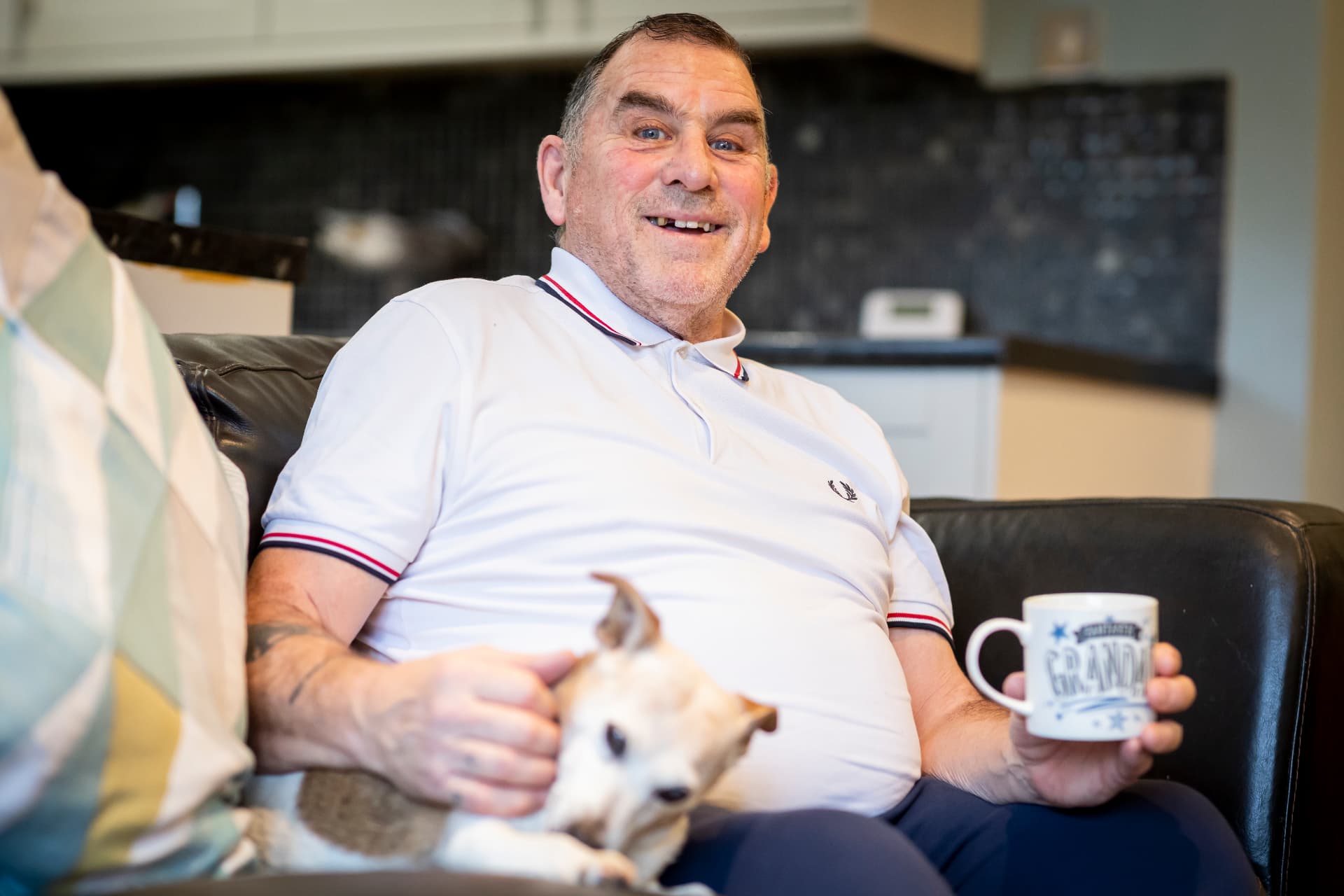 An older man managing at home on his sofa with his dog and a mug of tea